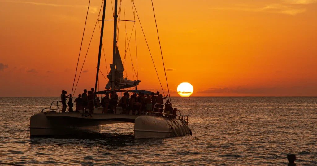 Sailboat catamaran on the ocean at sunset with a glowing orange sky, a popular romantic evening activity in Honolulu