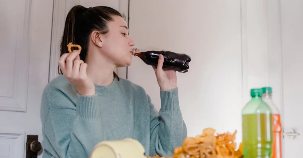 Woman drinking from a soda bottle at a table with snacks and drinks