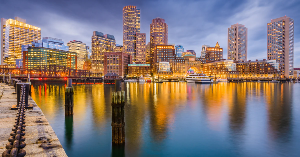 Boston skyline glowing at dusk with city lights reflecting on the harbor water